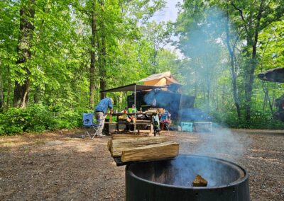 A view of a campsite with a close up shot of a fire ring and smoke. A CORE RV 5510 model in the background with a man and a child at the picnic table, a kayak mounted on a jeep to the right