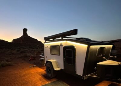 A nighttime shot of a white CORE RV 5510 model in the CO mountains at dusk with the sunset glowing the sky. The exterior perimeter lights are on and there is a place setting with chairs and a set of dog dishes
