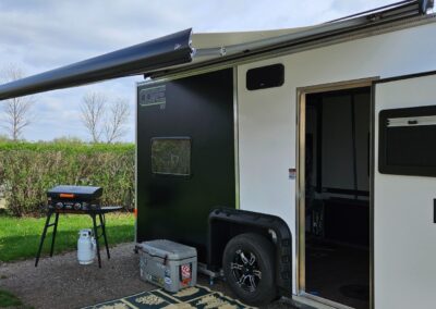 A close-up, outside view of a white and black CORE RV with the awning out and a cooler and Blackstone grill set outside