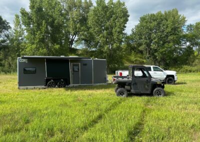 A ranger sitting in front of a white truck towing a grey and black CORE RV 8026 model with the awning out in a grassy field with trees in the background