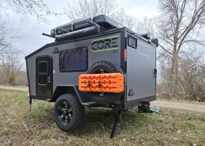 A grey CORE RV model with orange recovery boards strapped to the rear spare tire, sitting on the jacks with an outdoor shower and hard case rooftop tent on a dirt pathway