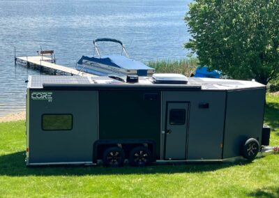 A CORE RV 8026 with solar panels and an awning set outside on the grass in front of a lake with a dock and a pontoon in the water