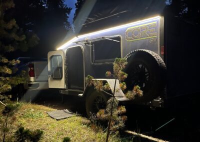 A nighttime shot of a CORE RV 5510 model in the woods with the door open, the window open and the exterior LED perimeter lights on highlighting the CORE RV decal