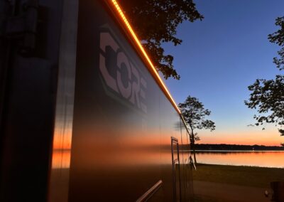 A night time shot of a CORE RV at the lake with a sunset glow and the exterior LED lights highlighting the CORE RV logo on the trailer